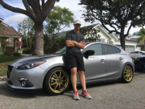 A middle-age man wearing a t-shirt, shorts, sneakers, and a baseball cap leans with arms crossed on a silver sports car in a suburban neighborhood.